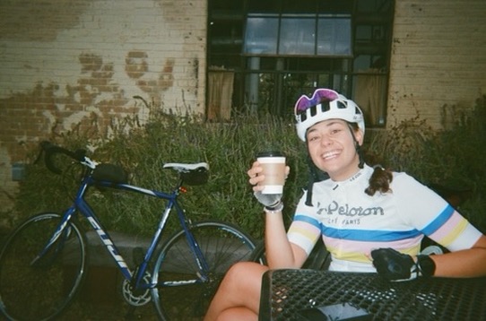 Maria with her bike, holding a coffee, wearing a Peloton Café Paris jersey